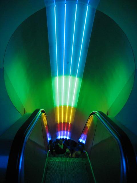 Looking down from the top of an escalator with rainbow lighting on the ceiling